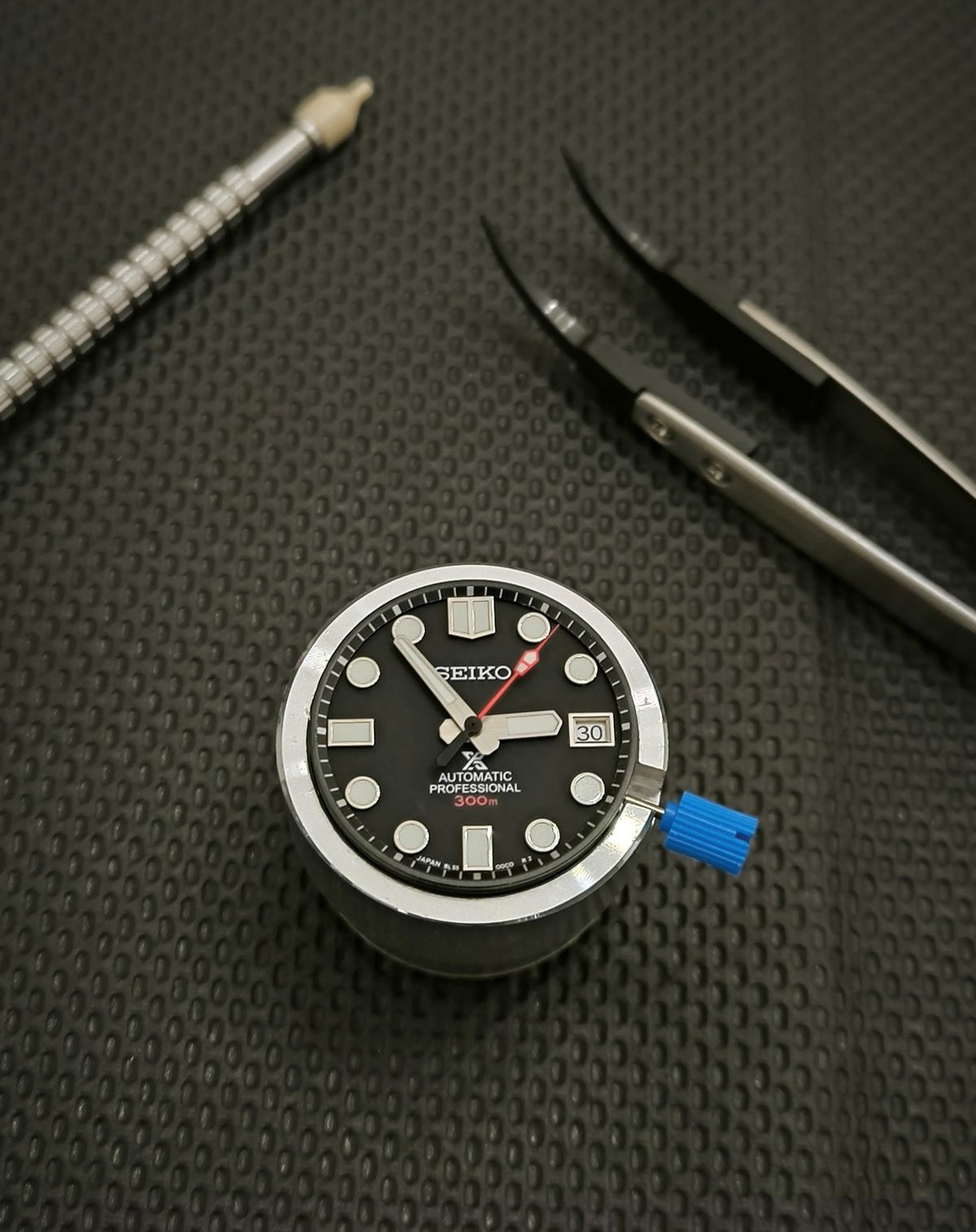 Close-up of a Seiko watch dial on a textured black surface with tools.